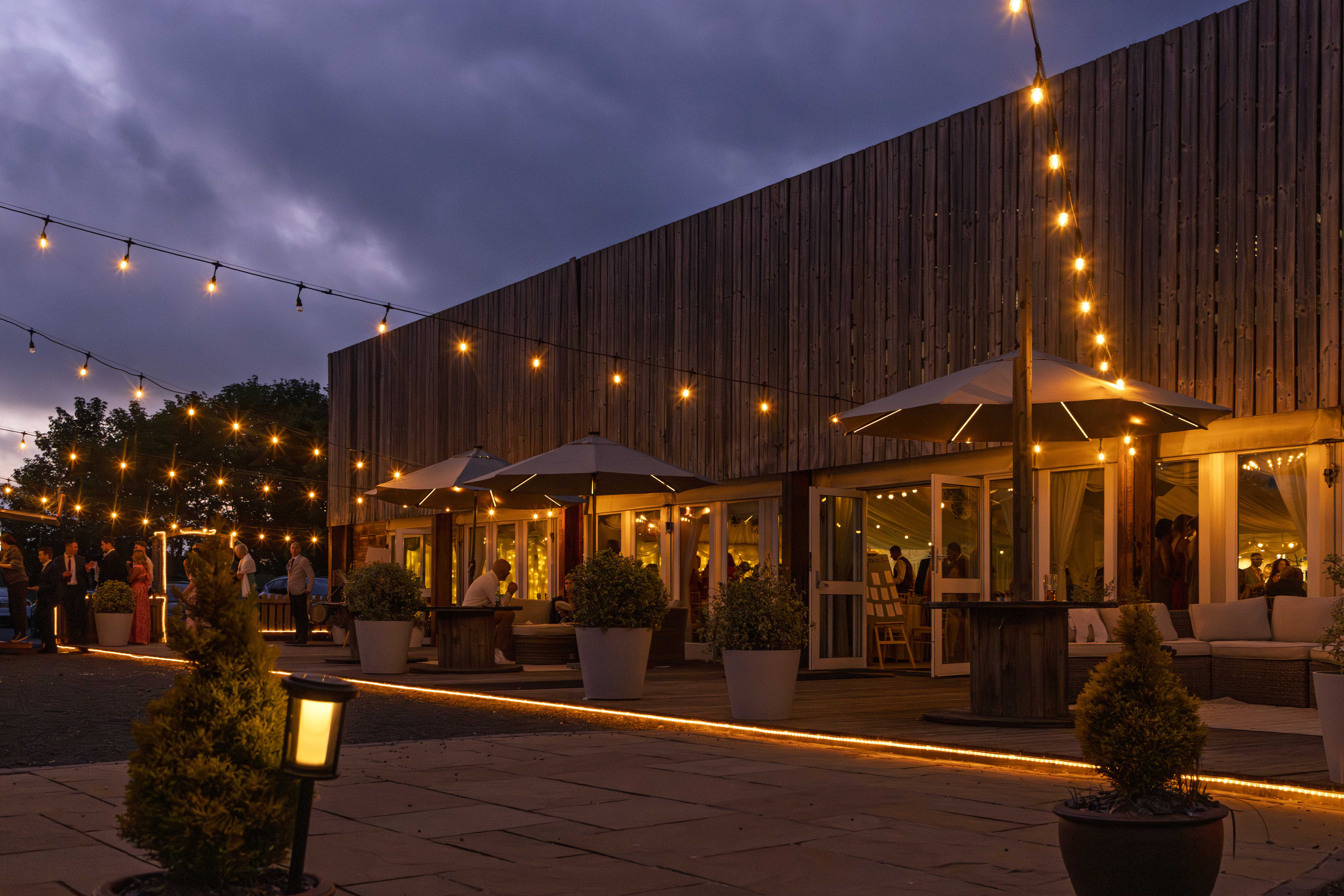 Guests mingling under glowing string lights during an outdoor evening reception at Vallum Weddings, with rustic wooden architecture, elegant umbrella seating, and a warm, inviting Northumberland countryside ambiance.