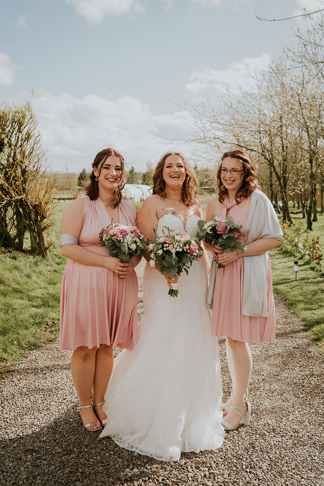 Bride in a white gown standing between two bridesmaids in matching blush dresses, all holding bouquets, posing on a countryside path at Vallum Weddings in Northumberland with lush greenery and rustic charm.